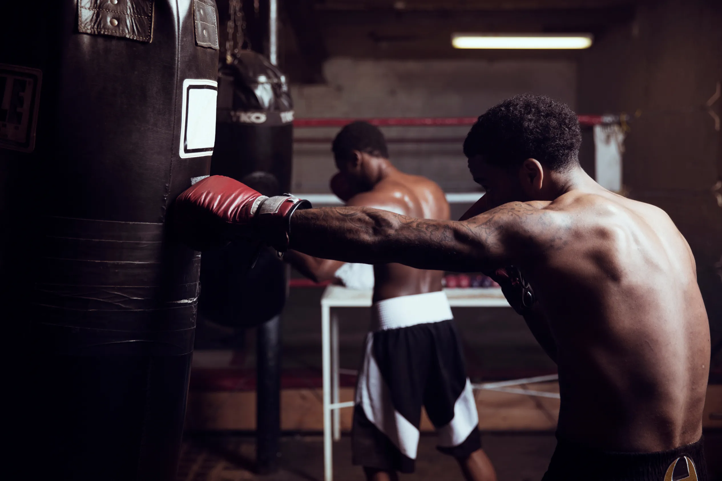 _Boxeur torse nu frappant un sac de frappe dans une salle sombre, concentré sur son entraînement pendant qu’un autre boxeur s’échauffe en arrière-plan._